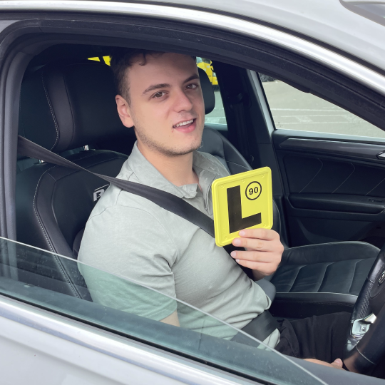 A man sitting in a grey car smiles proudly holding a yellow learners licence plate